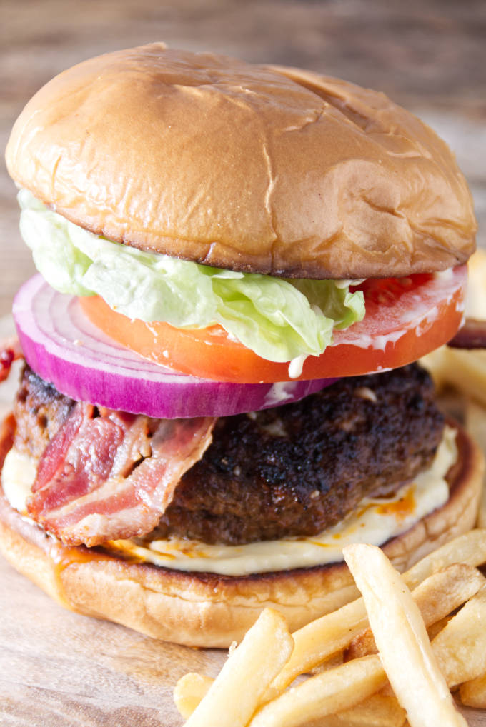 close up of a burger with bacon, onion, tomato, lettuce, and donkey sauce sitting on cutting board with fries