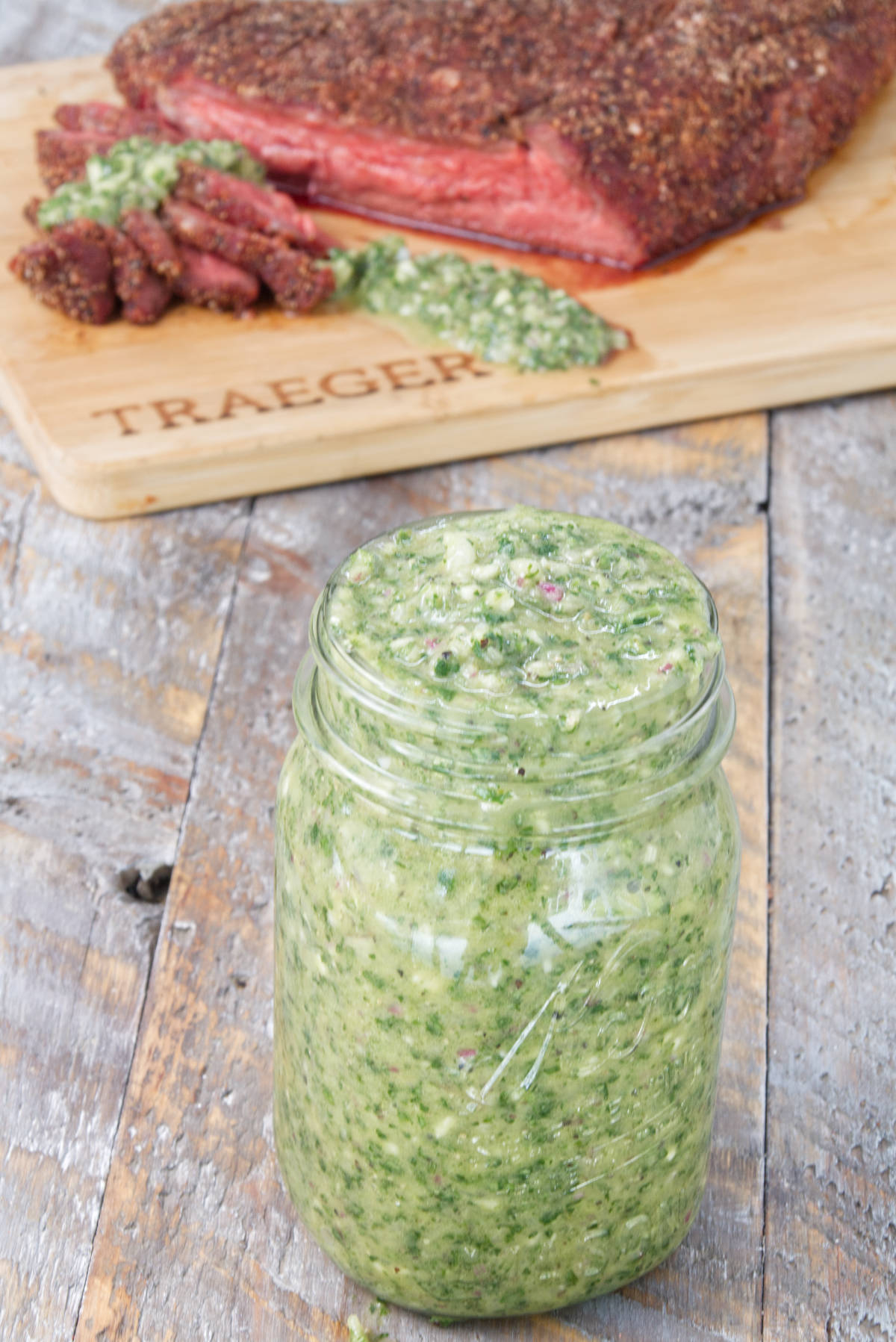 mason jar filled with vibrant green cilantro chimichurri sauce with sliced smoked tri-tip on the background on a cutting board