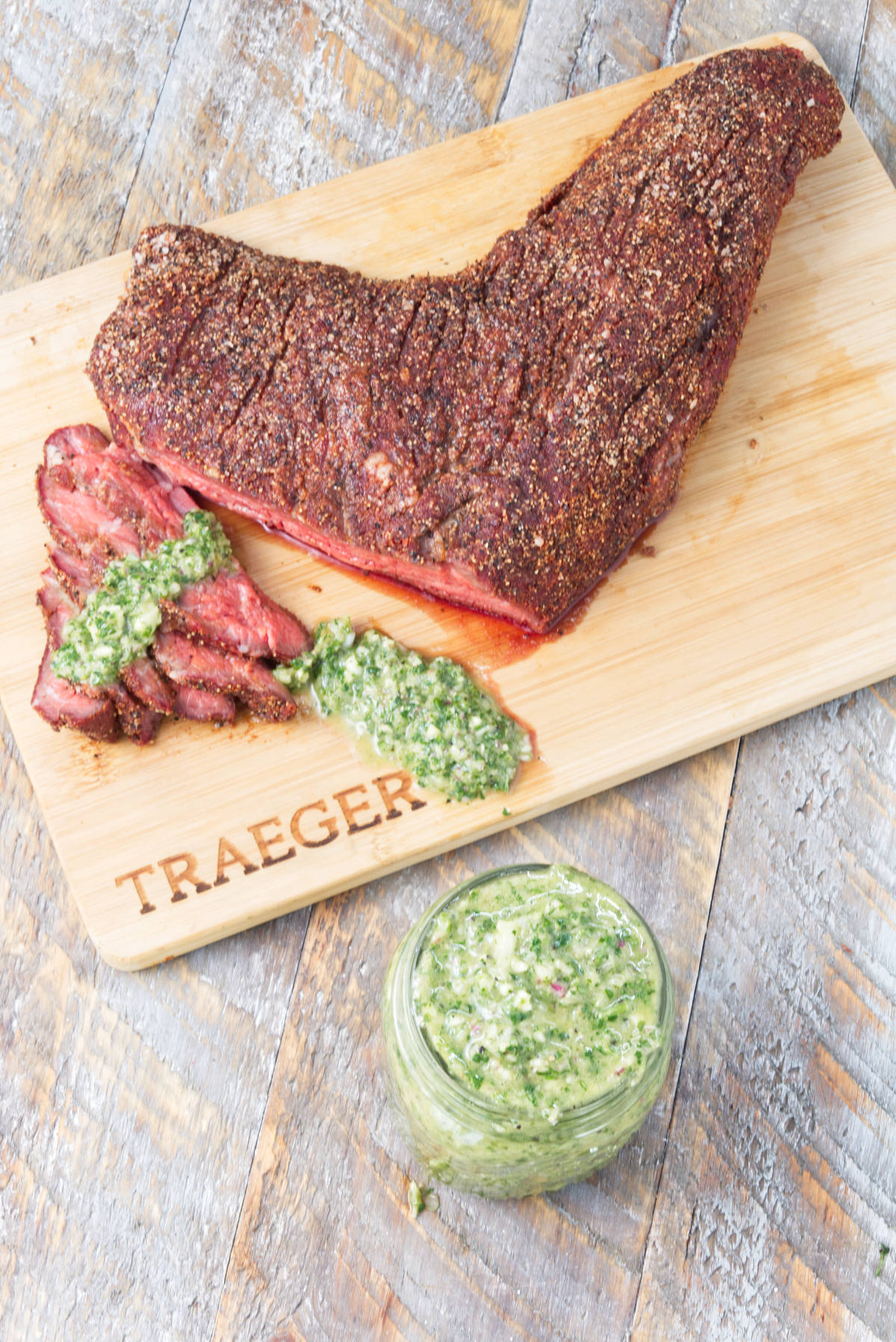 smoked tri-tip sliced on cutting board with cilantro chimichurri drizzled on top and chimichurri in mason jar