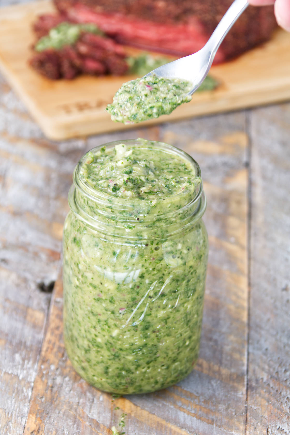 mason jar filled with vibrant green cilantro chimichurri sauce with sliced smoked tri-tip on the background on a cutting board