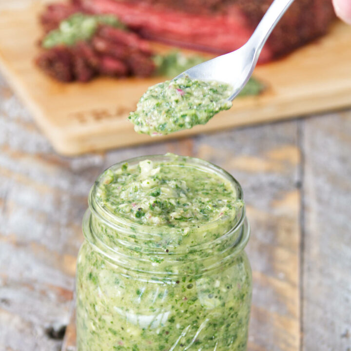 mason jar filled with vibrant green cilantro chimichurri sauce with sliced smoked tri-tip on the background on a cutting board