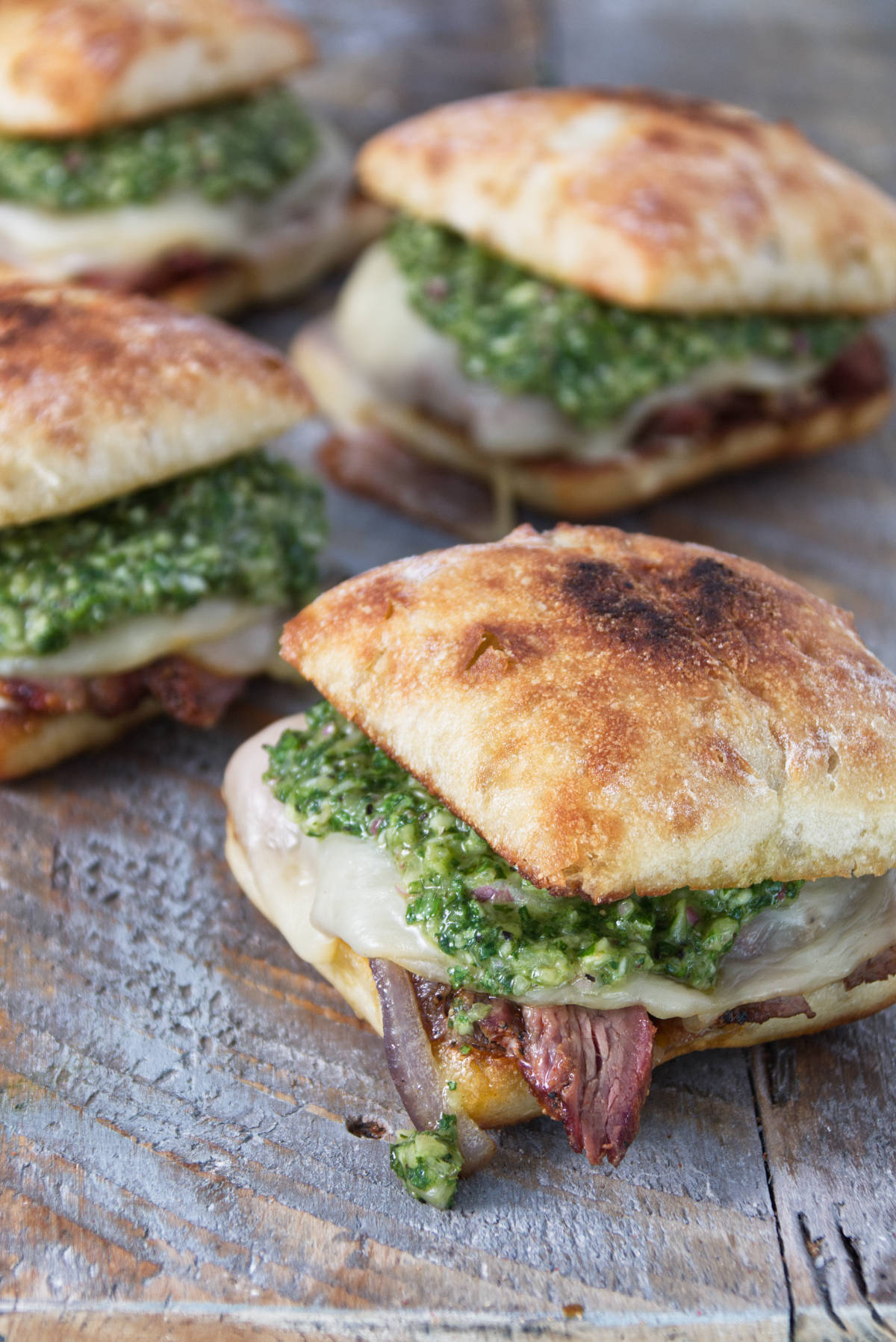 tri-tip sandwiches on a ciabatta roll with herby minto cilantro chimichurri and melted provolone cheese all on a wood cutting board