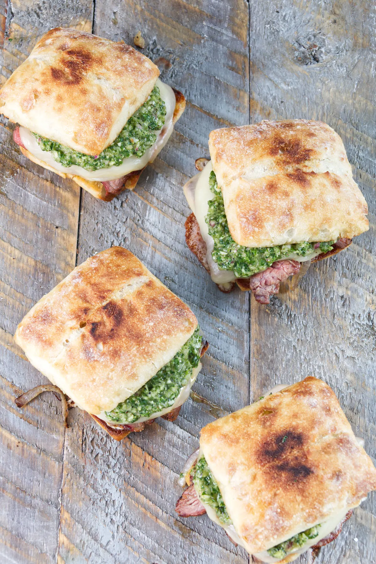 tri-tip sandwiches on a ciabatta roll with herby minto cilantro chimichurri and melted provolone cheese all on a wood cutting board