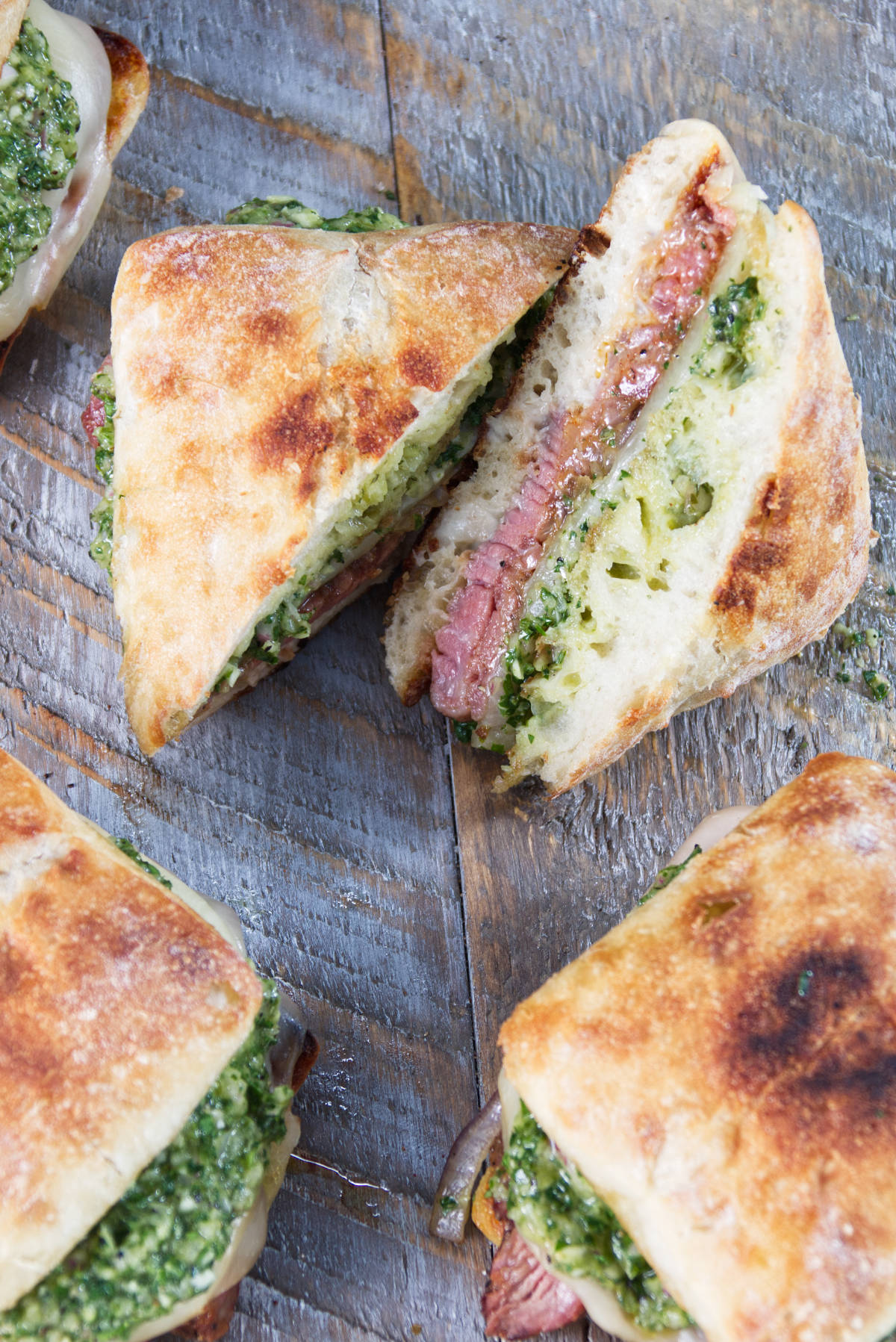 tri-tip sandwiches on a ciabatta roll with herby minto cilantro chimichurri and melted provolone cheese all on a wood cutting board