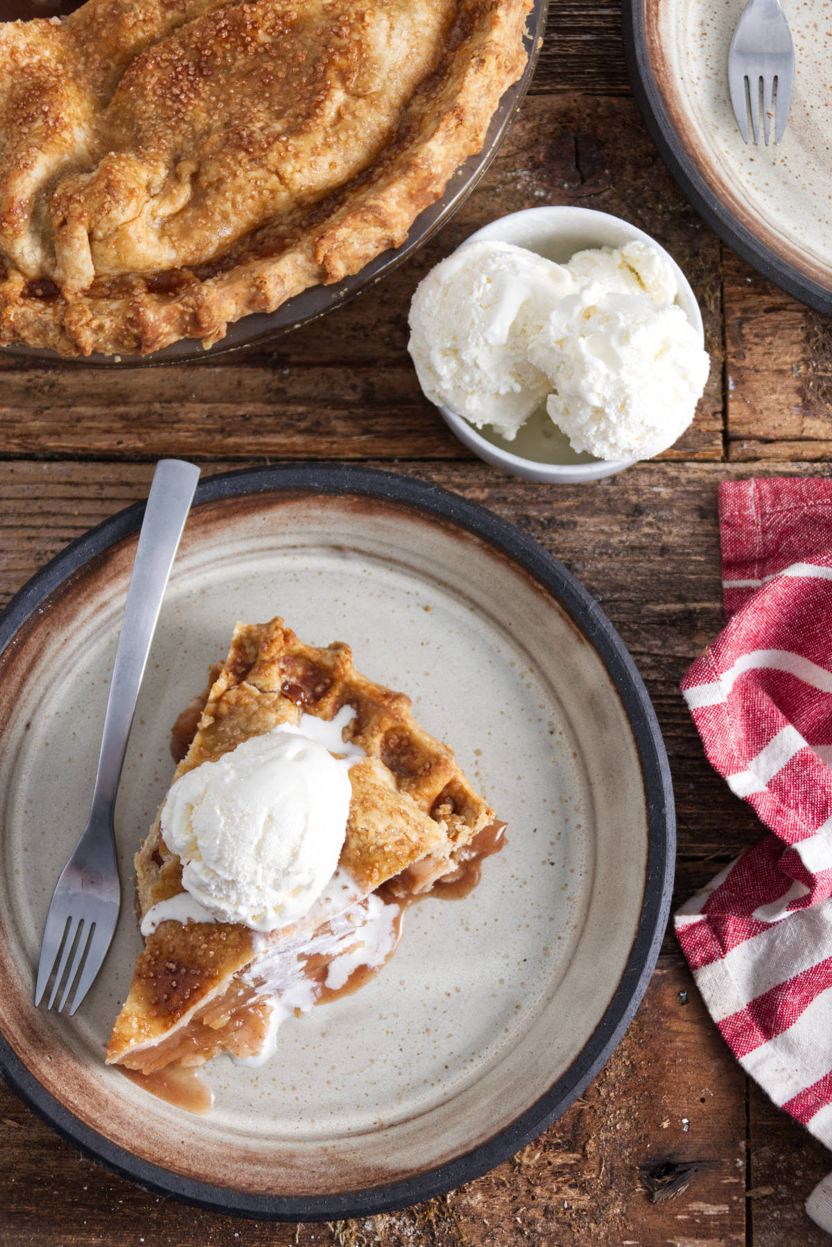 slice of apple pie on a white plate with a scoop of vanilla ice cream on top and bowl of ice cream and rest of pie