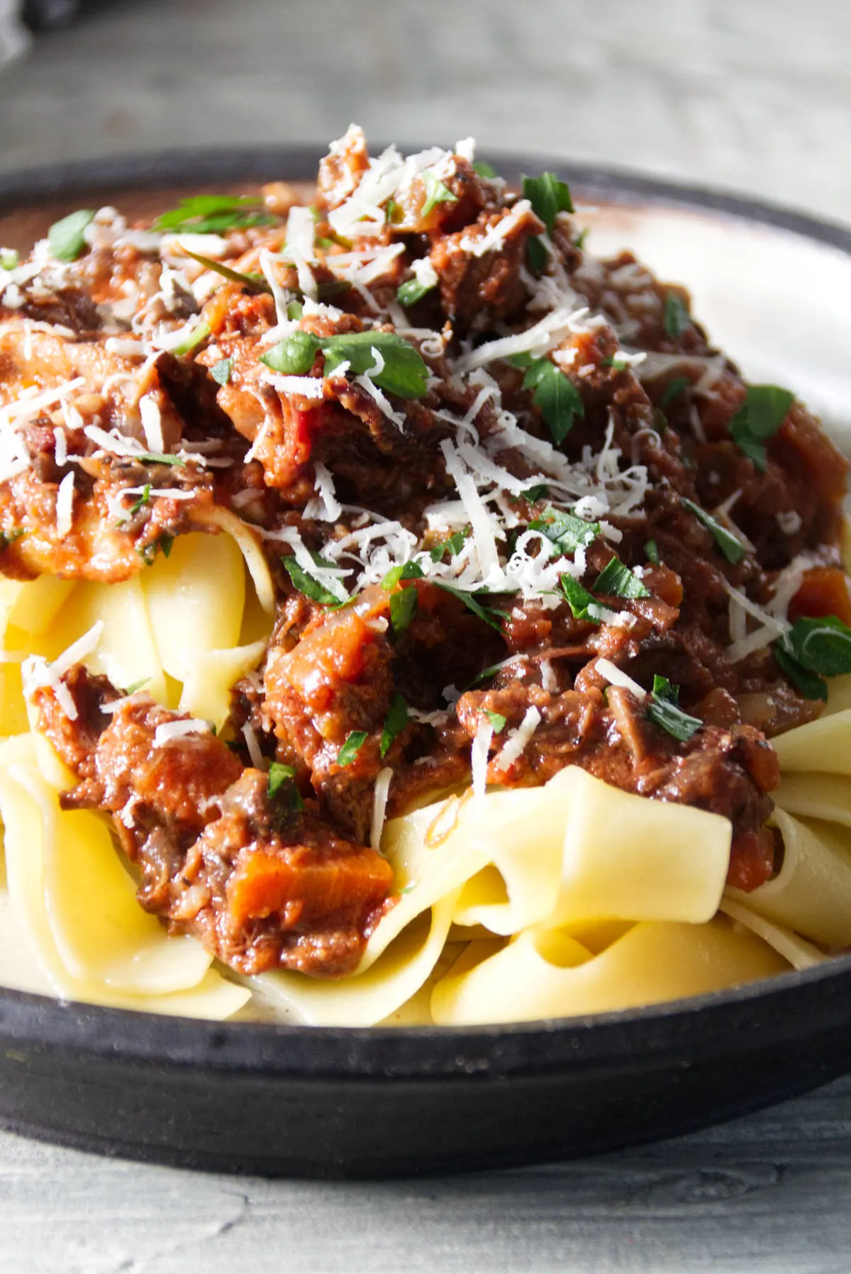 short rib ragu sauce served over pappardelle pasta on a plate and finished with grated parmesan and parsley.
