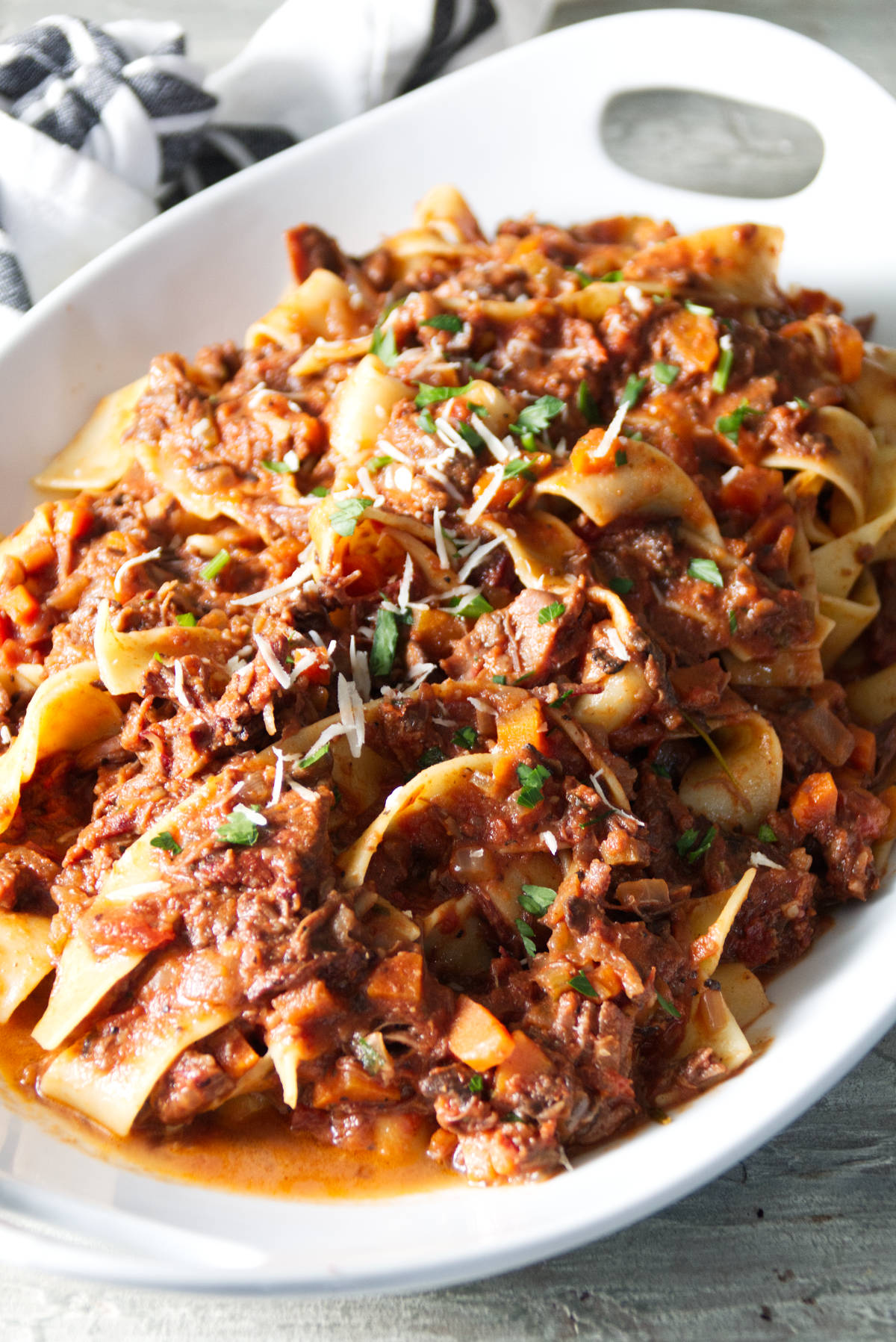 short rib ragu sauced served over pappardelle pasta in white bowl and finished with grated parmesan and parsley.