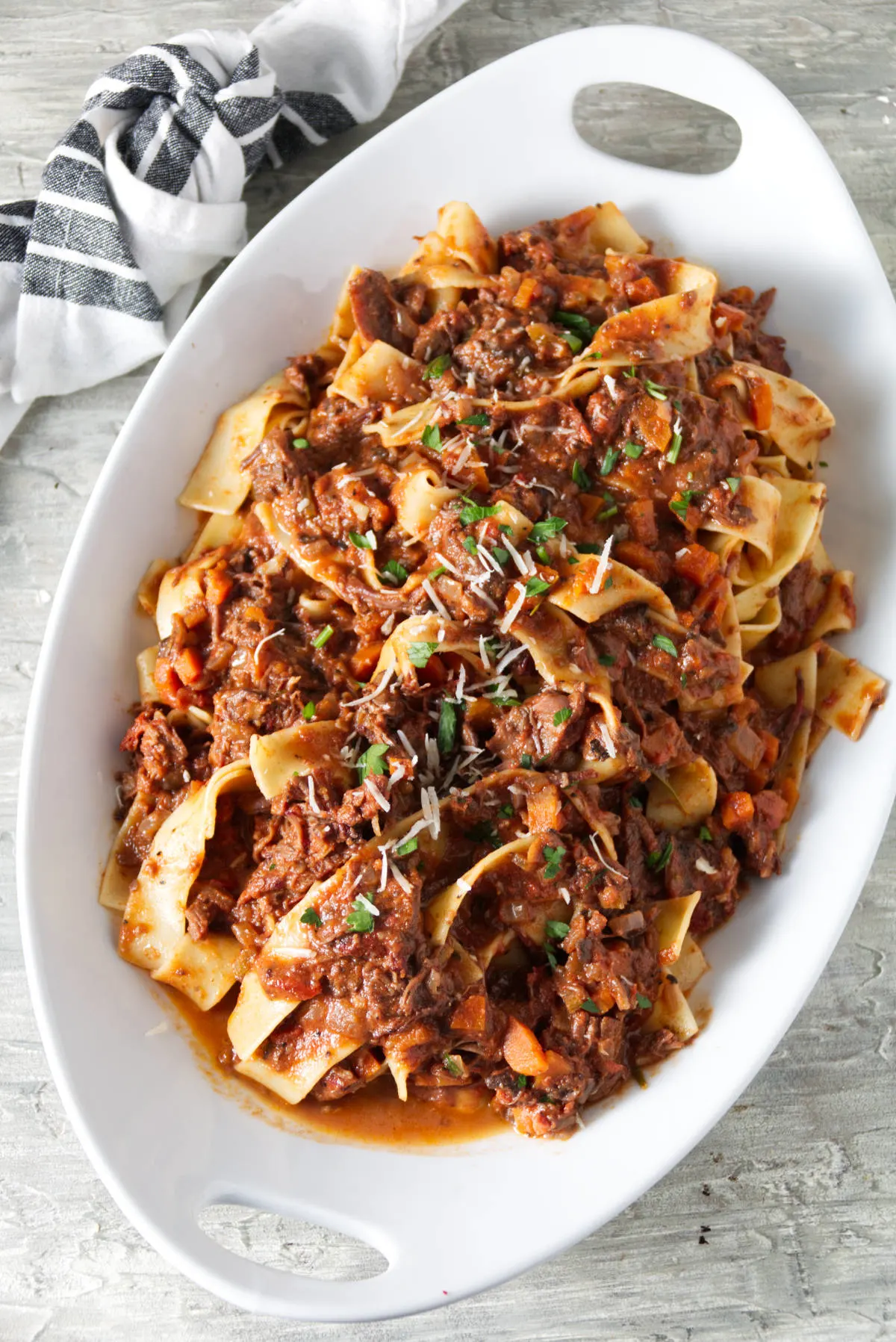 short rib ragu sauced served over pappardelle pasta in white bowl and finished with grated parmesan and parsley.