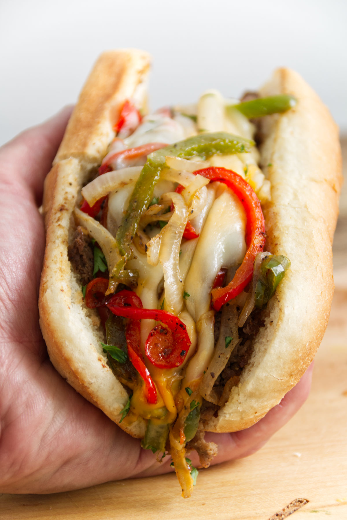 philly cheesesteak sandwich being held in hand and topped with onions peppers and gooey cheese whiz and provolone