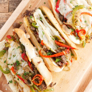 three philly cheesesteak sandwiches laid out on cutting board, each one filled with steak, red and green peppers, melted provolone, and cheese whiz