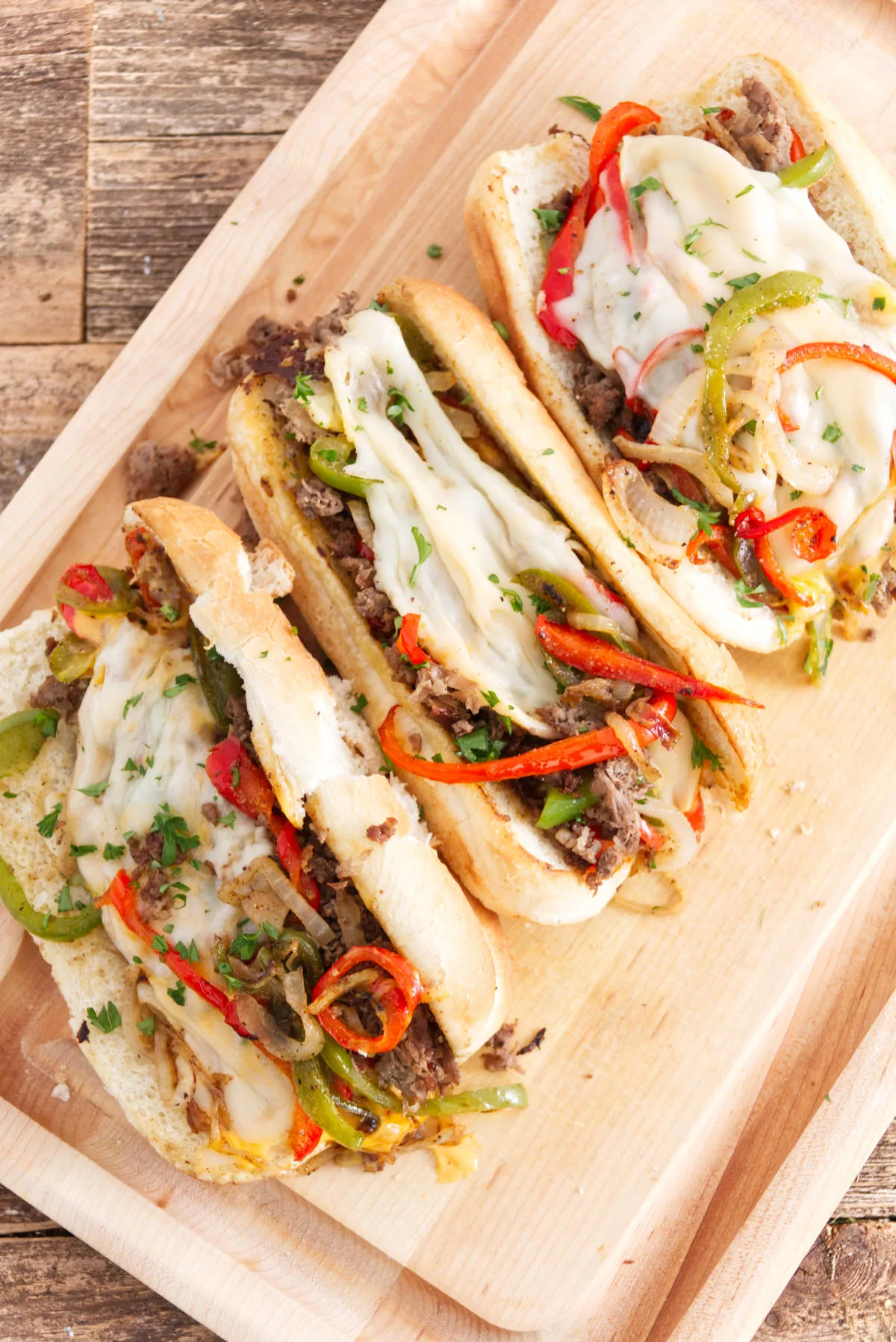 three philly cheesesteak sandwiches laid out on cutting board, each one filled with steak, red and green peppers, melted provolone, and cheese whiz