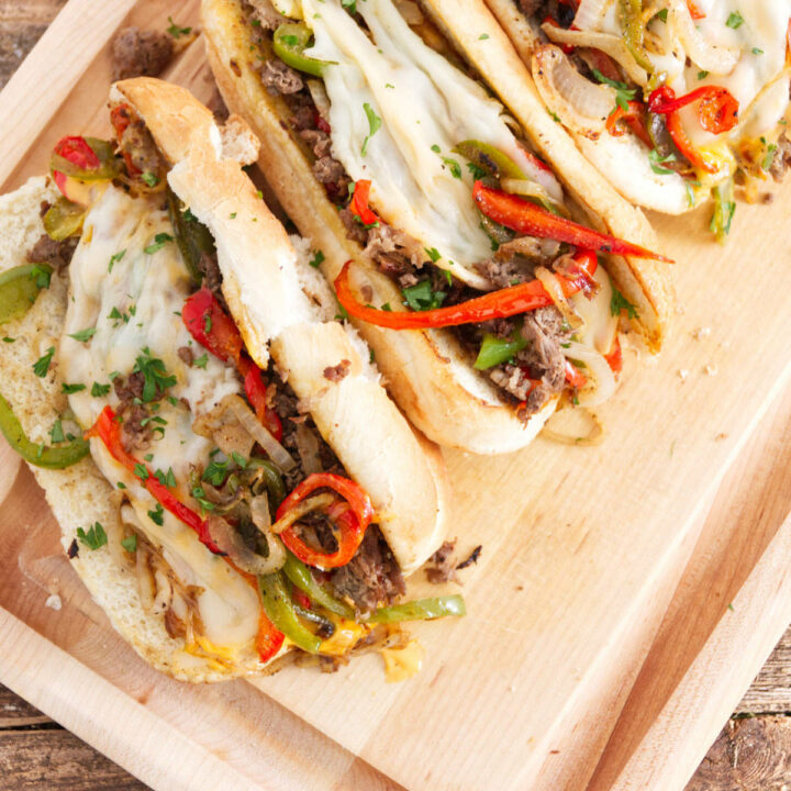 three philly cheesesteak sandwiches laid out on cutting board, each one filled with steak, red and green peppers, melted provolone, and cheese whiz