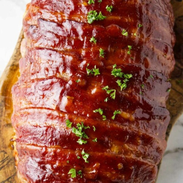 Beautifully carmelized bacon wrapped meatloaf resting on a wood cutting board.