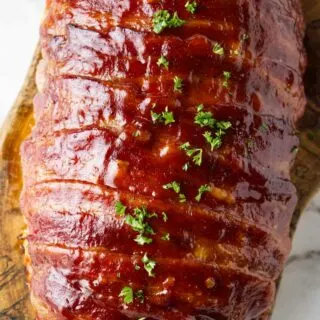 Beautifully carmelized bacon wrapped meatloaf resting on a wood cutting board.