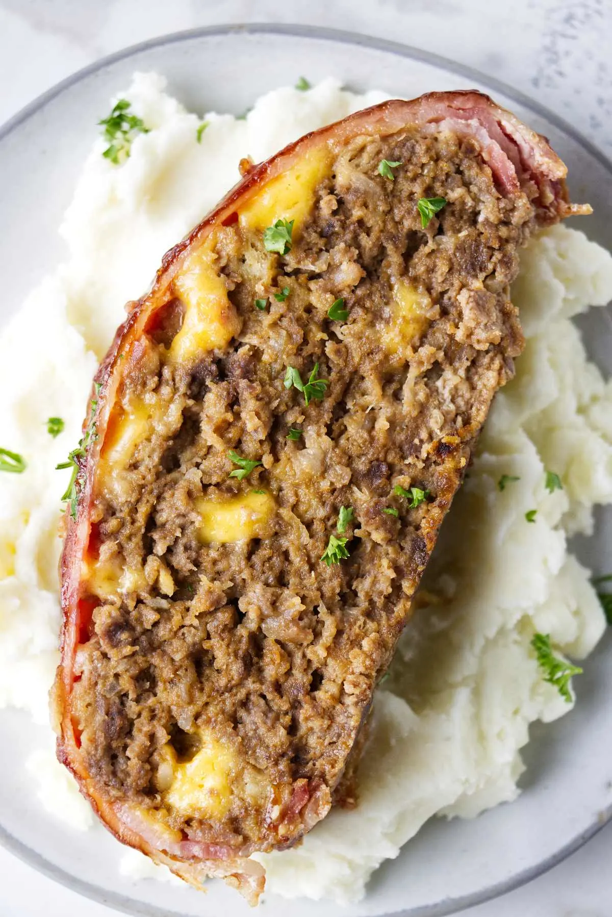 A slice of bacon wrapped meatloaf sitting on a bed of mashed potatoes.