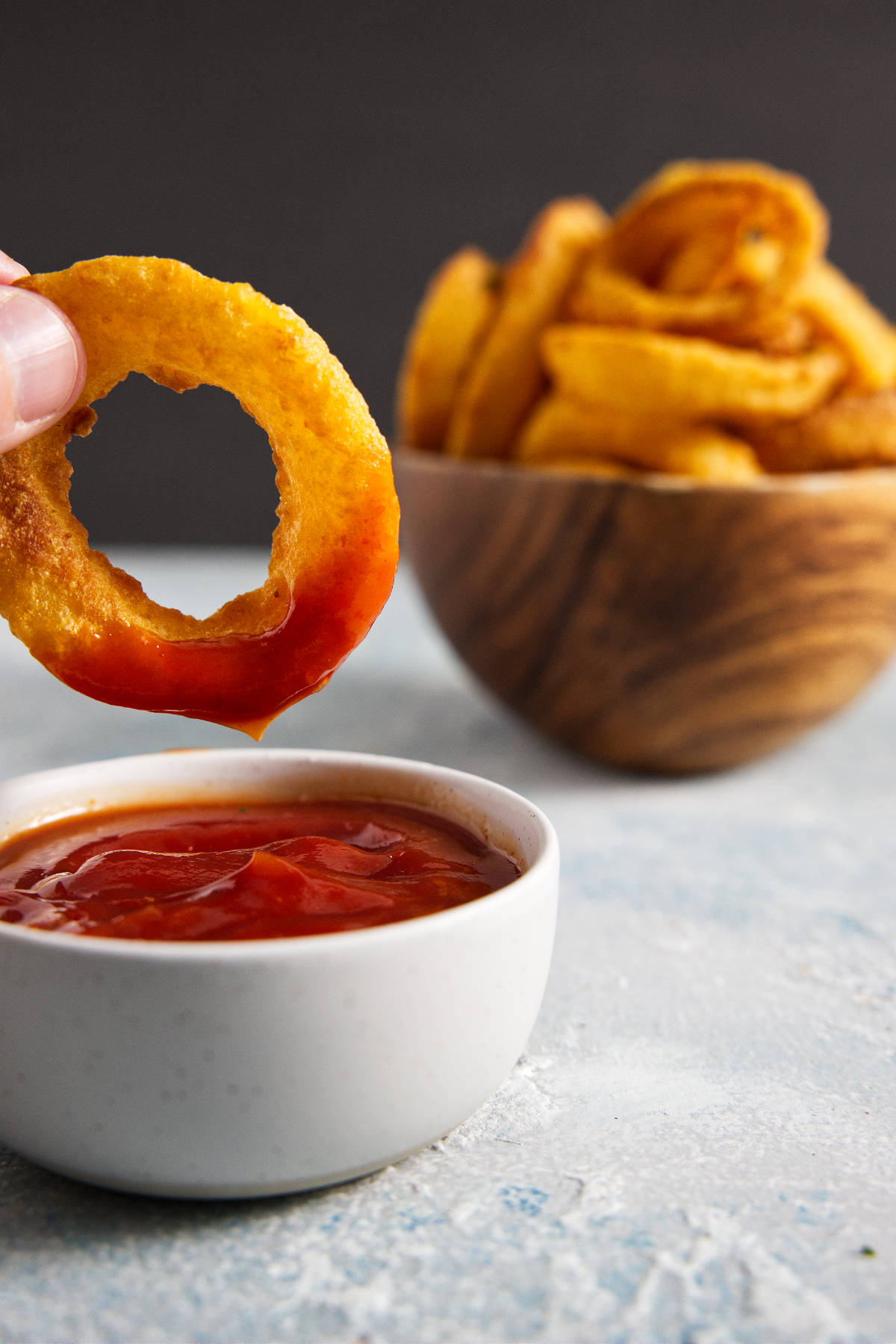 Onion ring being dipped into ketchup with wooden bowl of onion rings in background