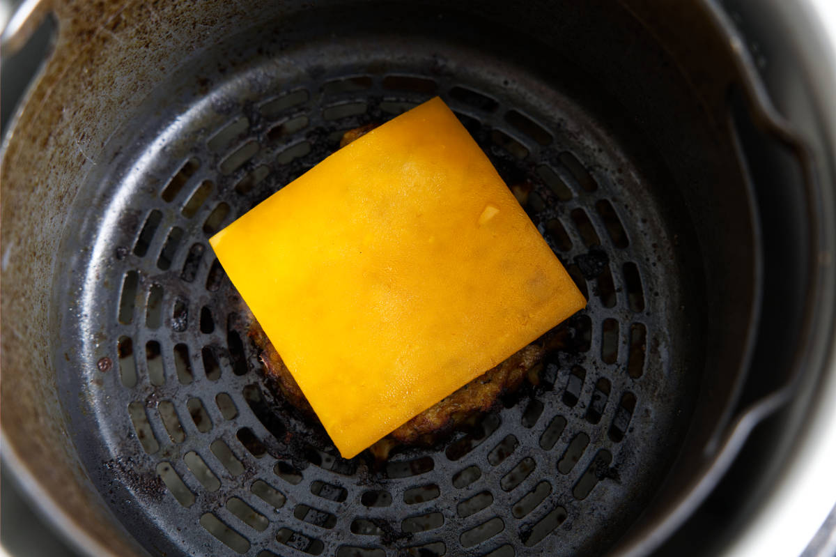 Melting slice of cheddar cheese on top of turkey burger patty in air fryer.