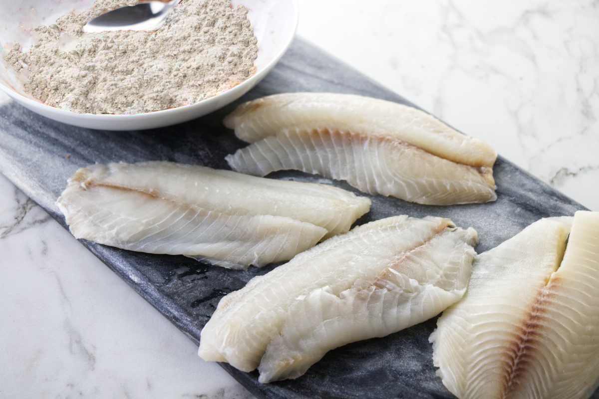 Fresh tilapia fish filets next to a bowl of flour.