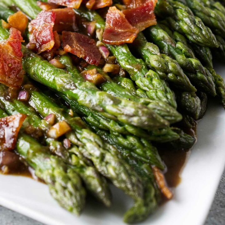 Cooked asparagus on white plate with chopped bacon and onion vinaigrette.