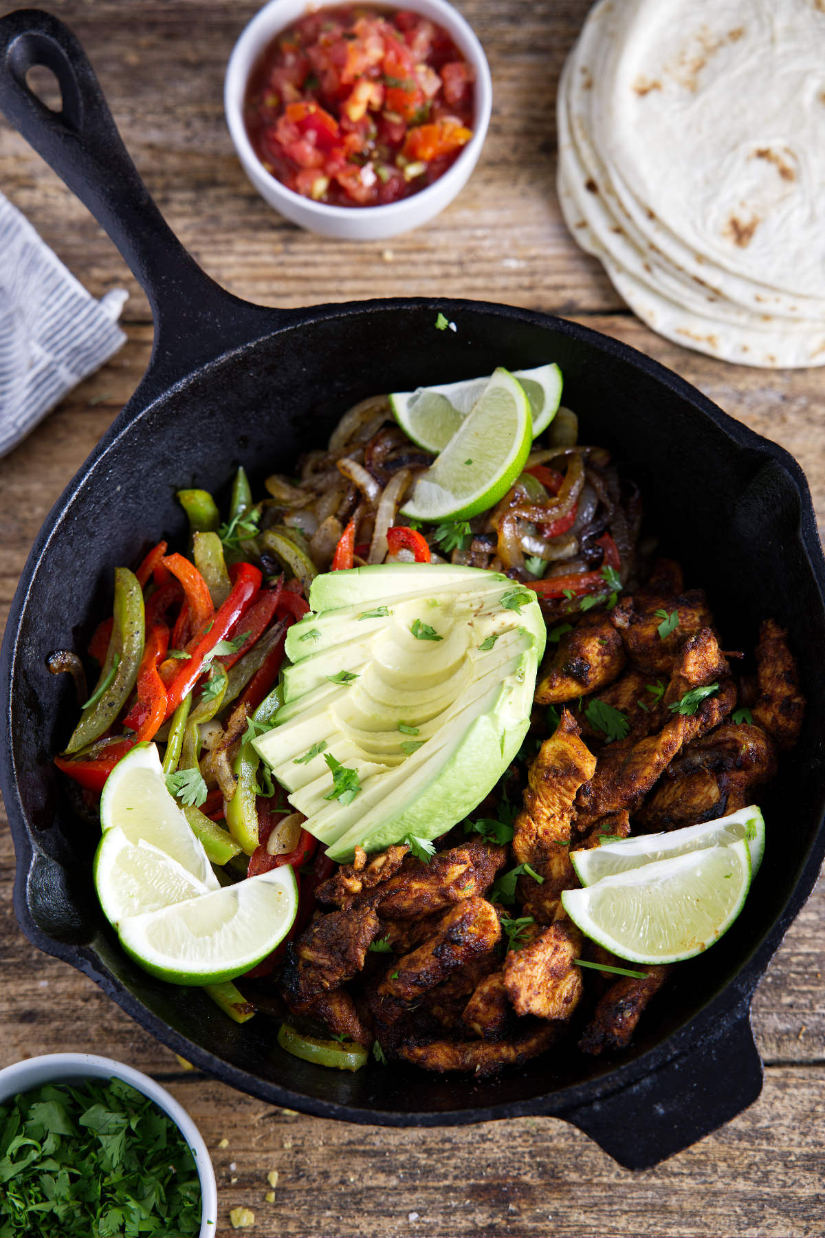 Cast iron filled with chicken fajitas that have been carmelized to perfection and topped with limes and avocado and surrounded by tortillas, salsa, and cilantro.