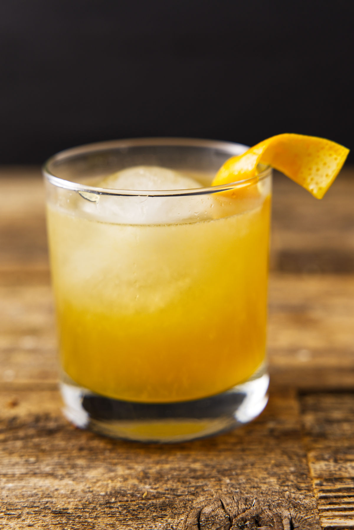 Glass filled with orange bourbon cocktail with orange peel twist and globe ice.