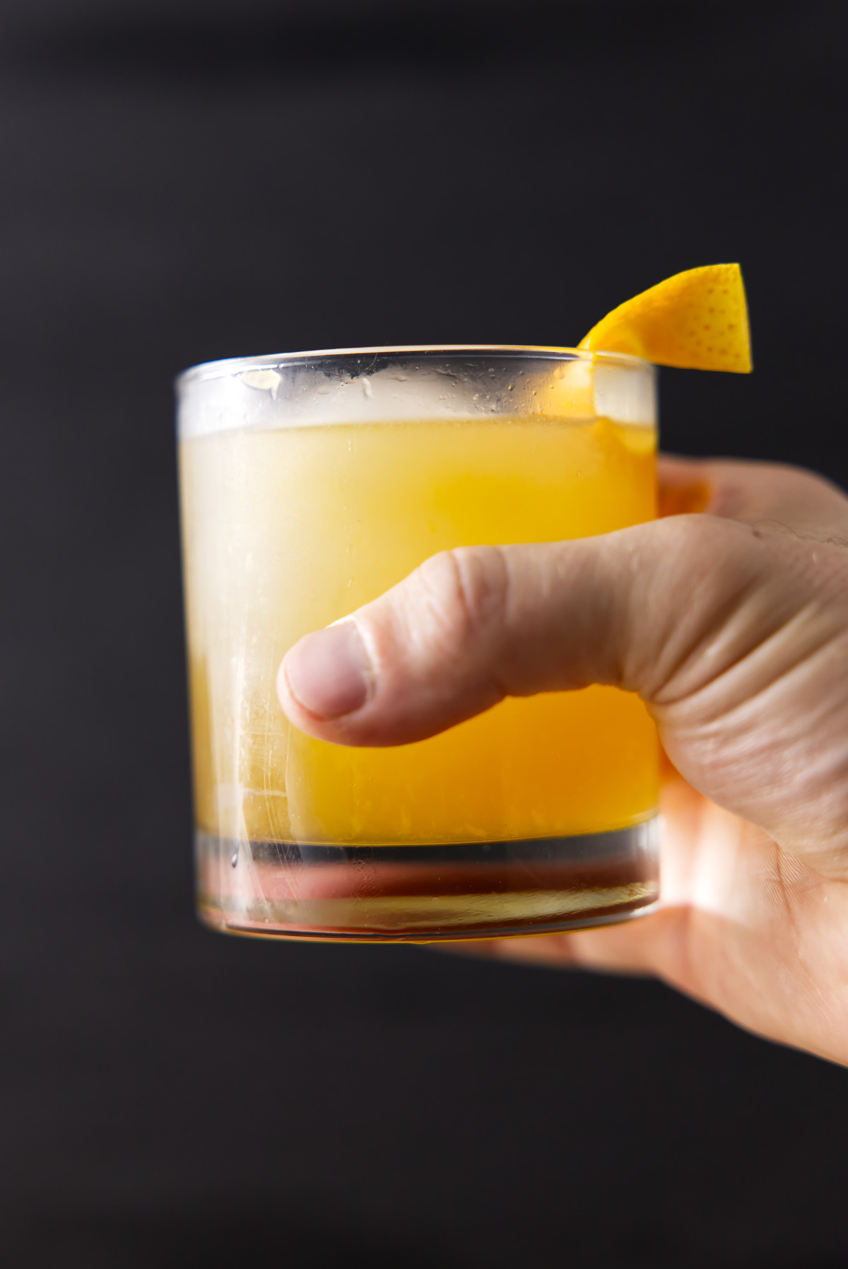 Hand holding glass filled with maple bourbon smash cocktail with orange twist.