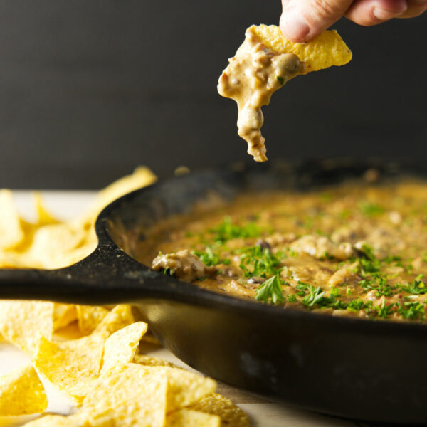 Gooey cheese dripping from chip with skillet of smoked cheese dip underneath.
