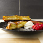Pouring syrup over a stack of stuffed French toast with whipped cream and raspberries.