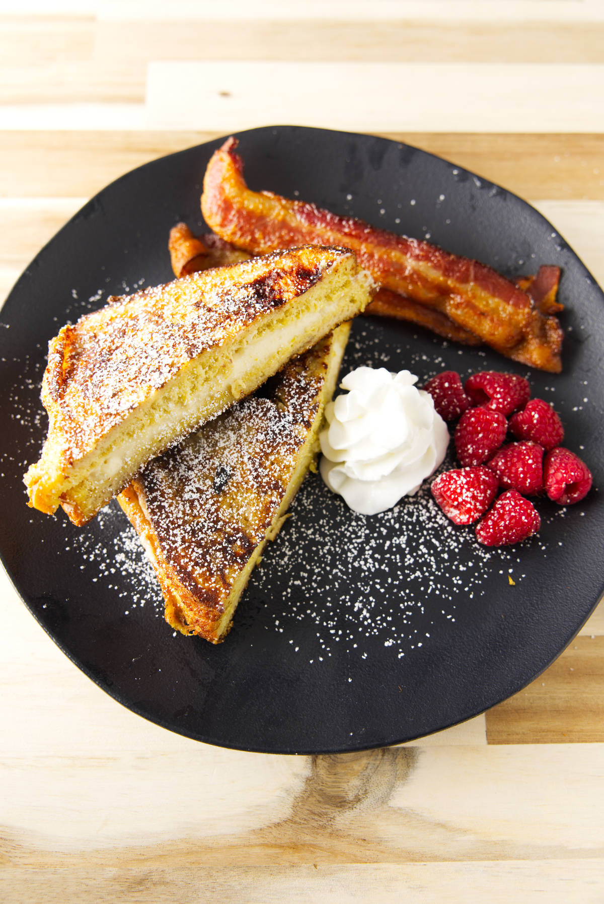 Stuffed French toast plated with bacon, raspberries, and whipped cream and dusted with powdered sugar.