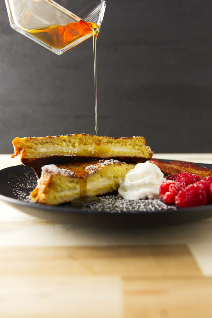 Pouring syrup over a stack of stuffed French toast with whipped cream and raspberries.