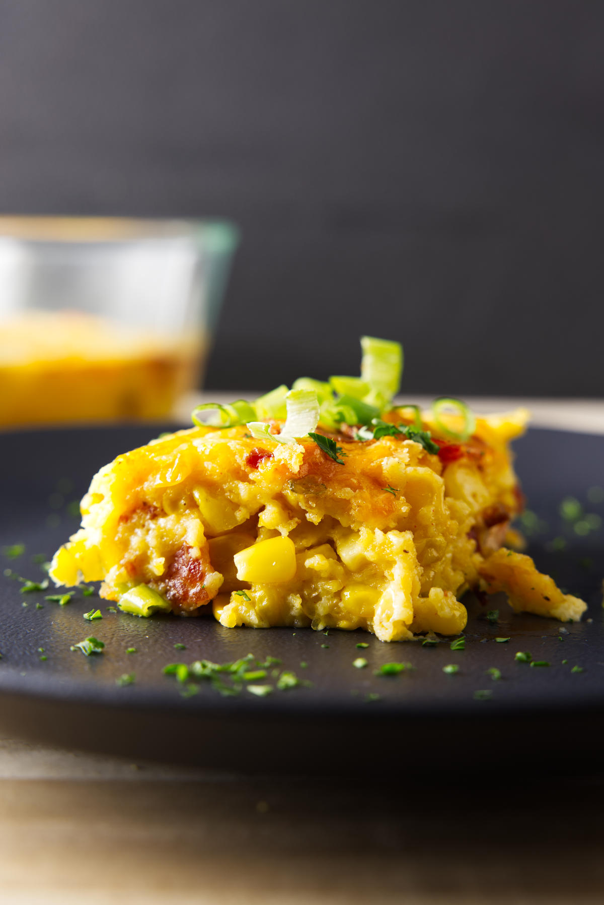 Close up shot of a scoop of Smoked Corn Casserole on a black plate with pan in the back.