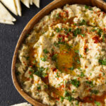 Close up of smoked baba ganoush in wooden bowl topped with paprika, chopped parsley, and an olive oil drizzle.