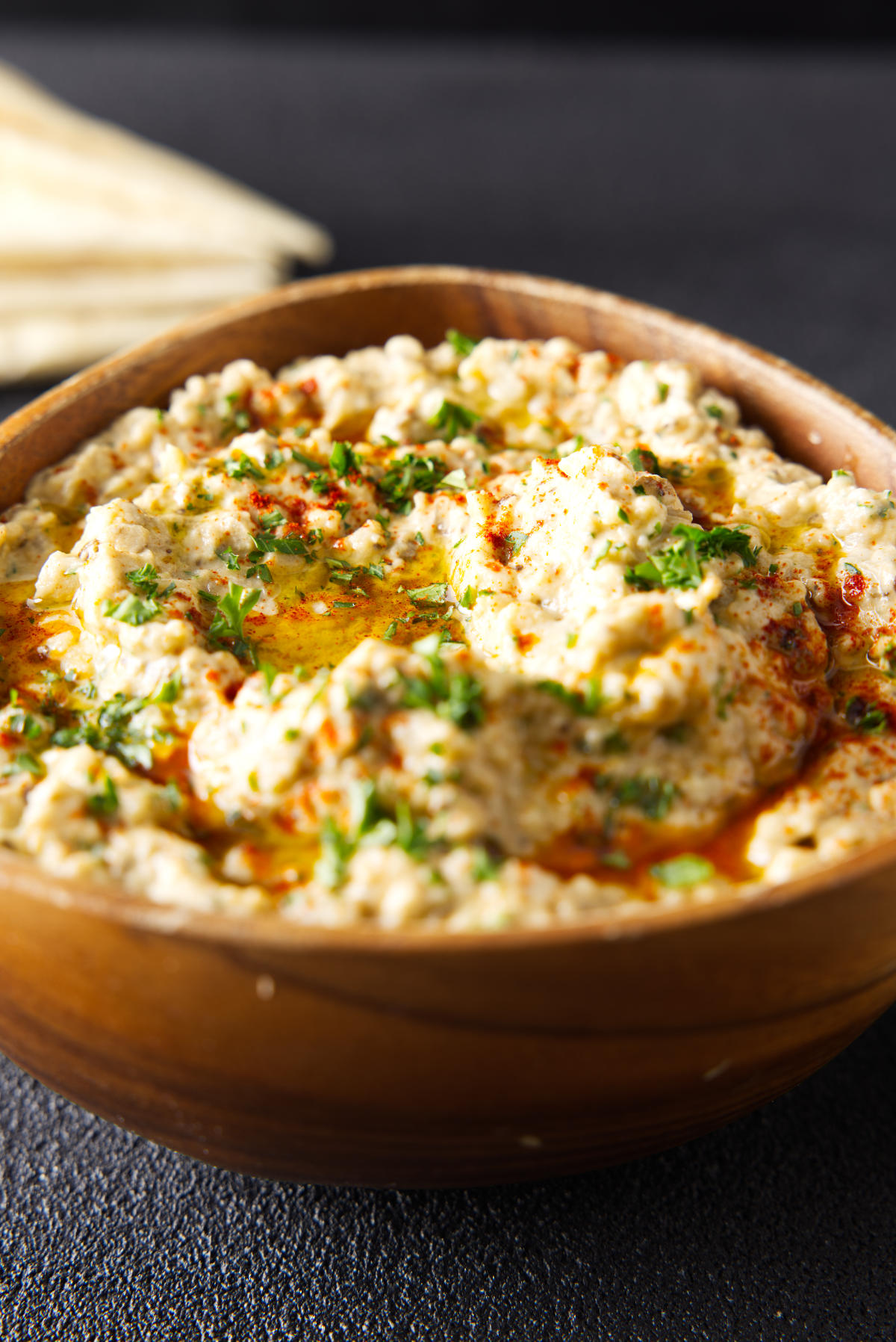 Wooden bowl filled to the brim with baba ganoush and topped with a drizzle of olive oil and chopped parsley.