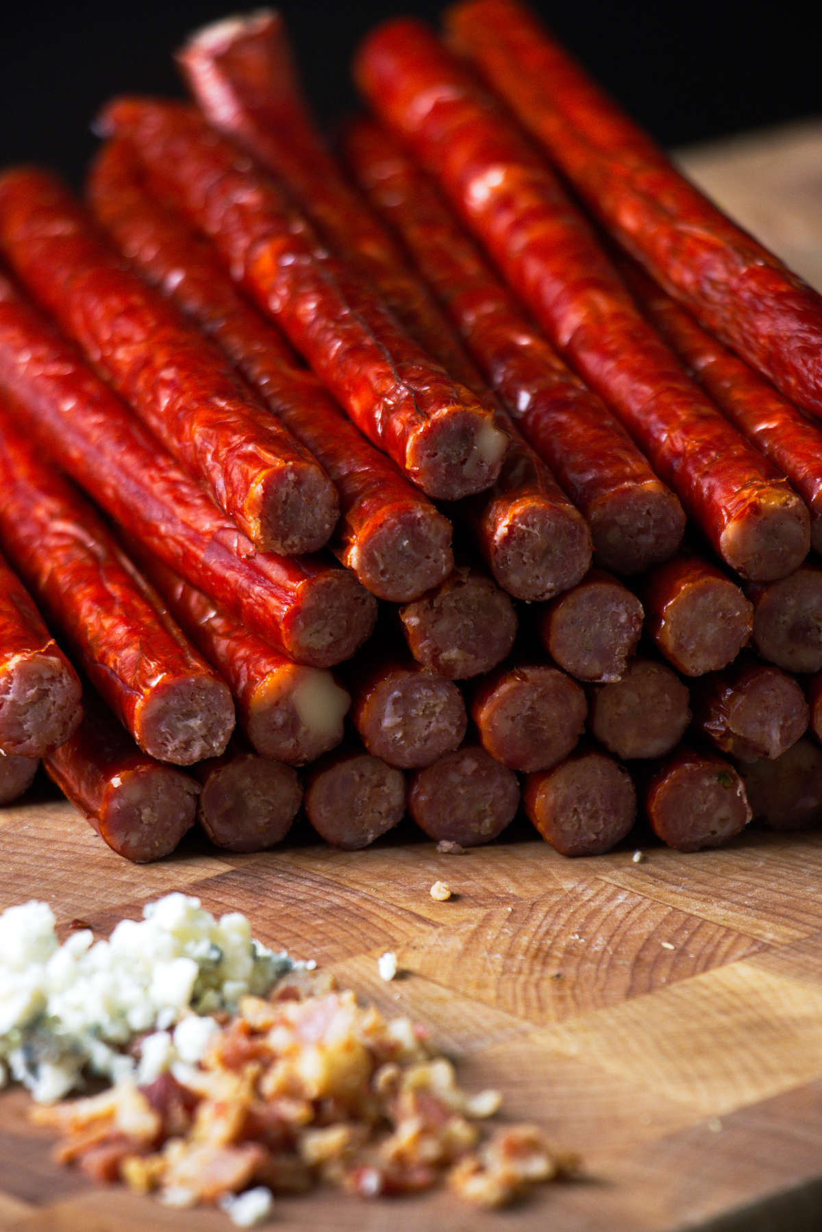 Blue cheese bacon venison snack sticks stacked on a butcher block with blue cheese and bacon crumbles.