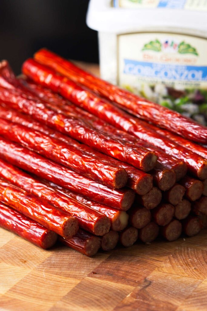 Snack sticks stacked on butcher block with gorgonzola cheese in background.