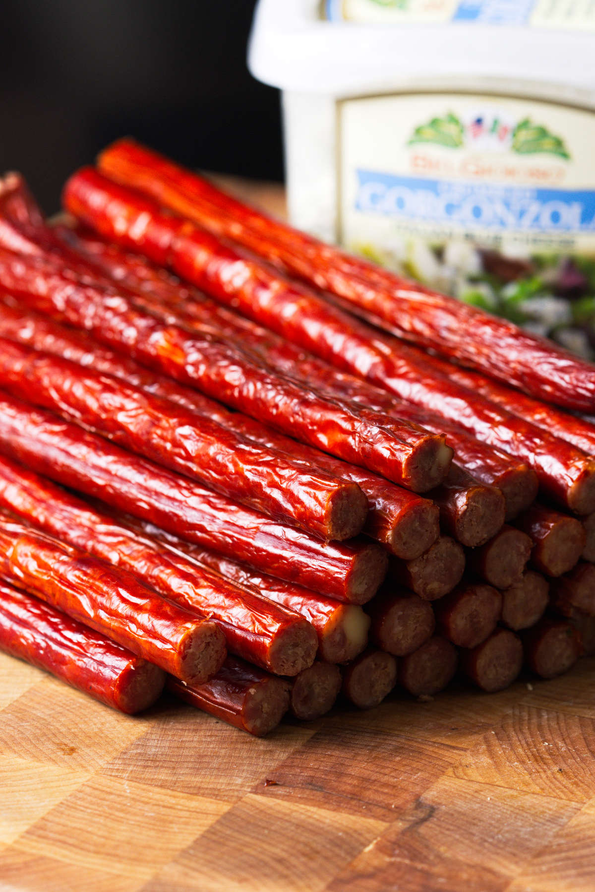Snack sticks stacked on butcher block with gorgonzola cheese in background.