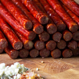 Close up of blue cheese venison snack sticks on butcher block with blue cheese crumbles and bacon in foreground.