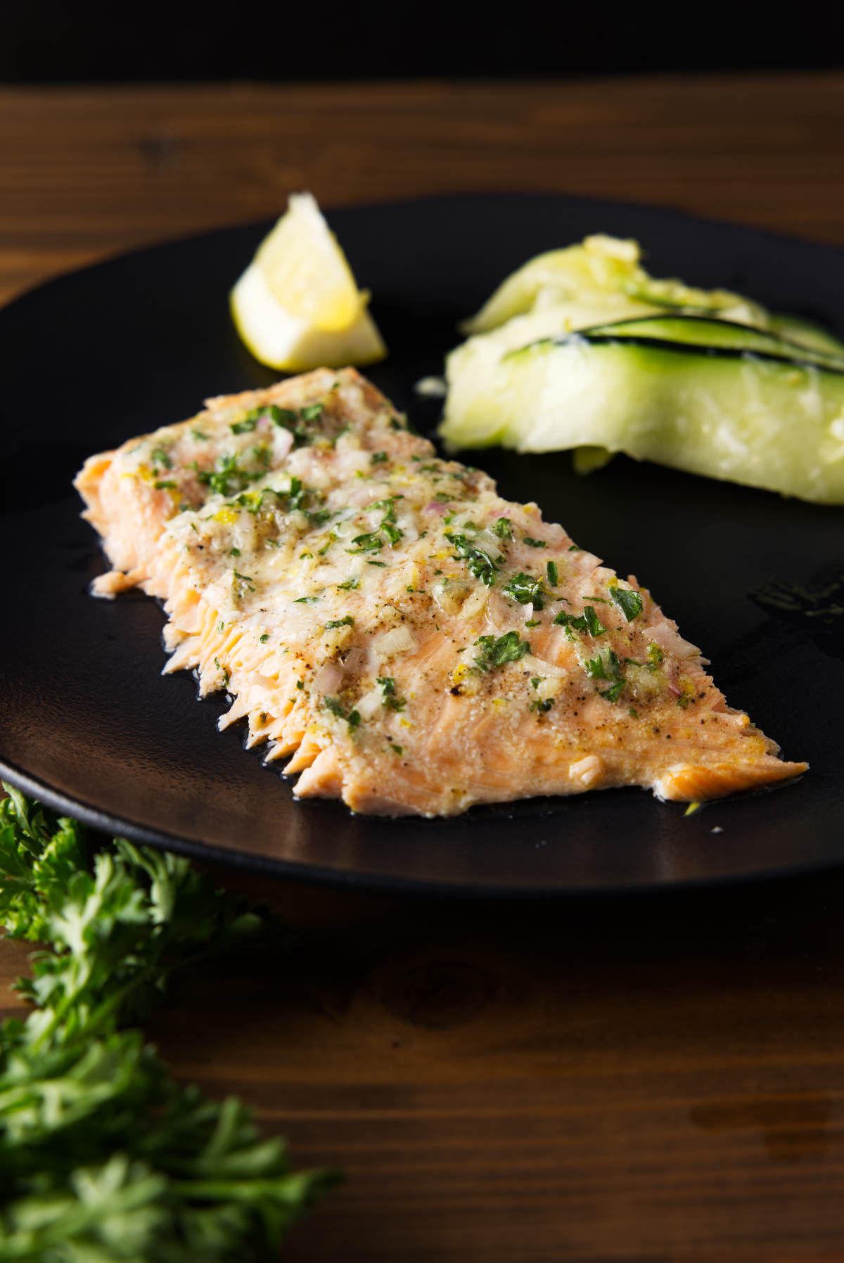 Slice of salmon with compound butter on top served on a black plate with a lemon wedge and zucchini salad.