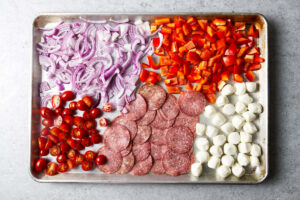Spreading sliced onions, tomatoes, salami, bell peppers, and mozzarella out on a sheet pan to smoke.