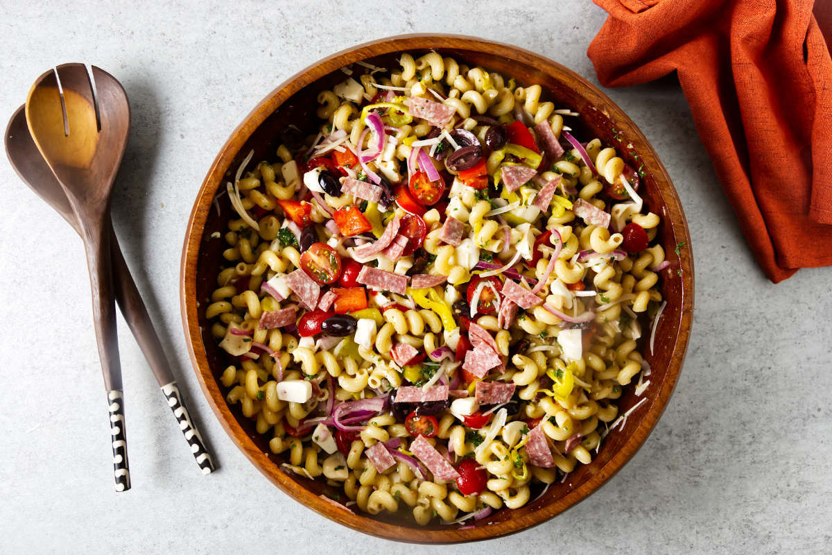 A wooden salad bowl filled with Italian pasta salad next to salad utensils.