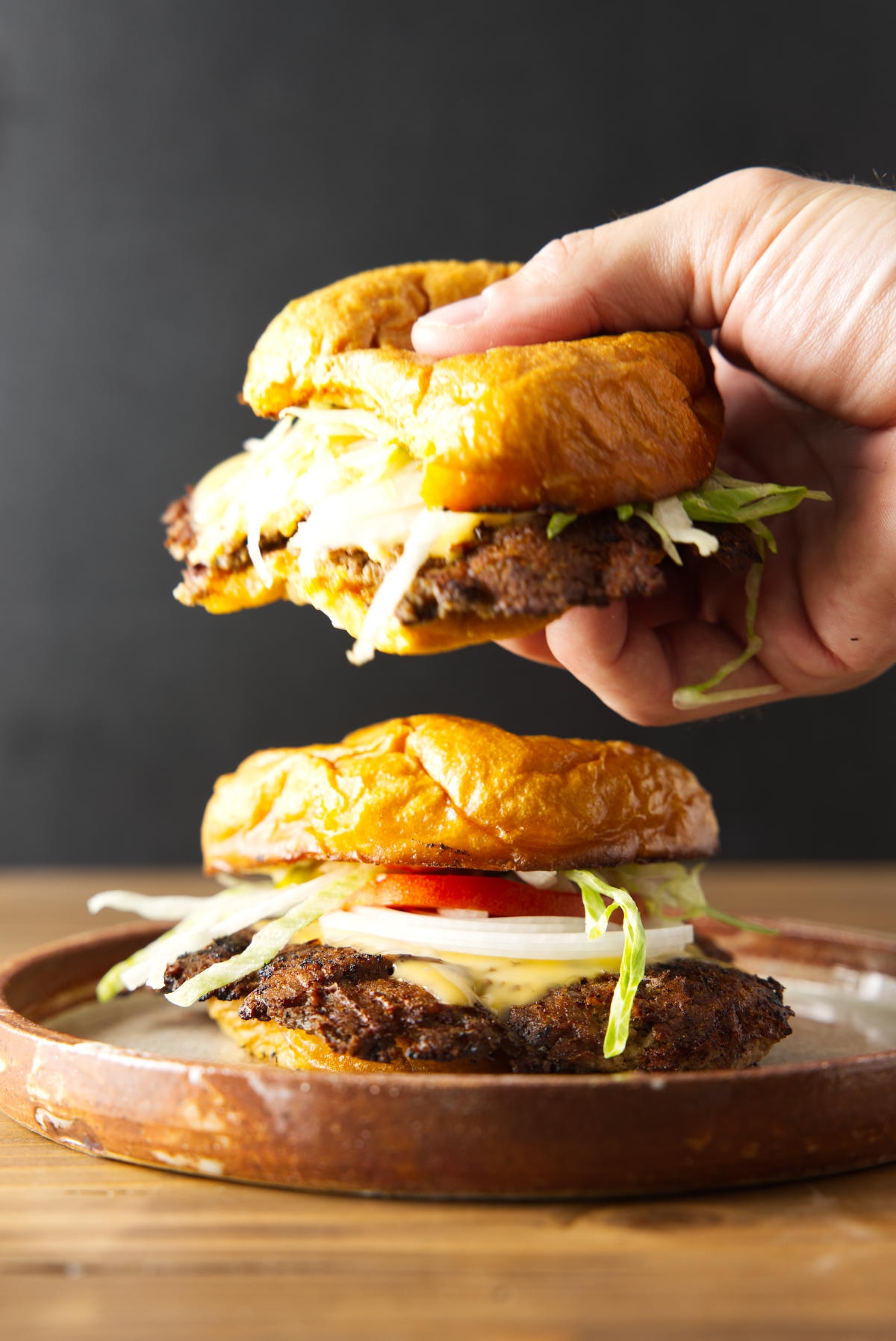 Holding a smash burger hovering over another plated smash burger loaded with toppings.