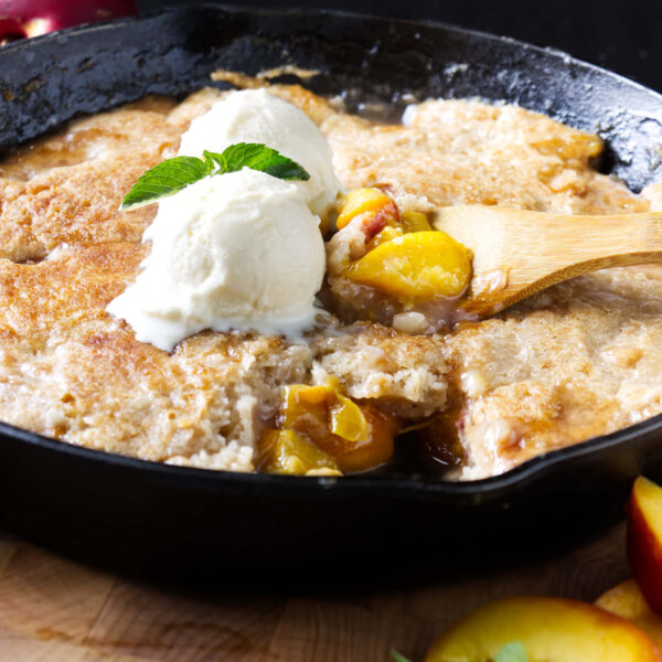 Scooping smoked peach cobbler from a cast iron skillet.