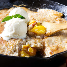 A spoon scooping peach cobbler from a cast iron skillet with vanilla ice cream.