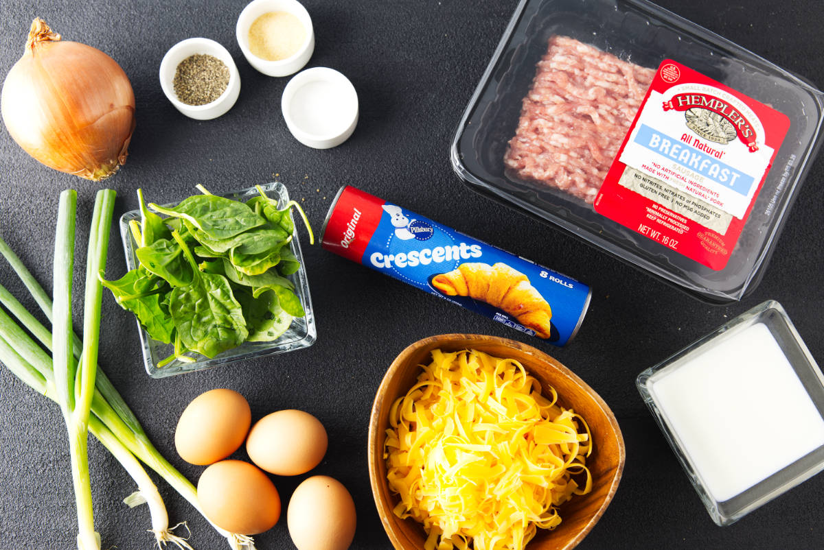 Crescent roll dough, breakfast sausage, eggs, milk, shredded cheese, spinach, onions, and seasonings arranged on a dark counter.