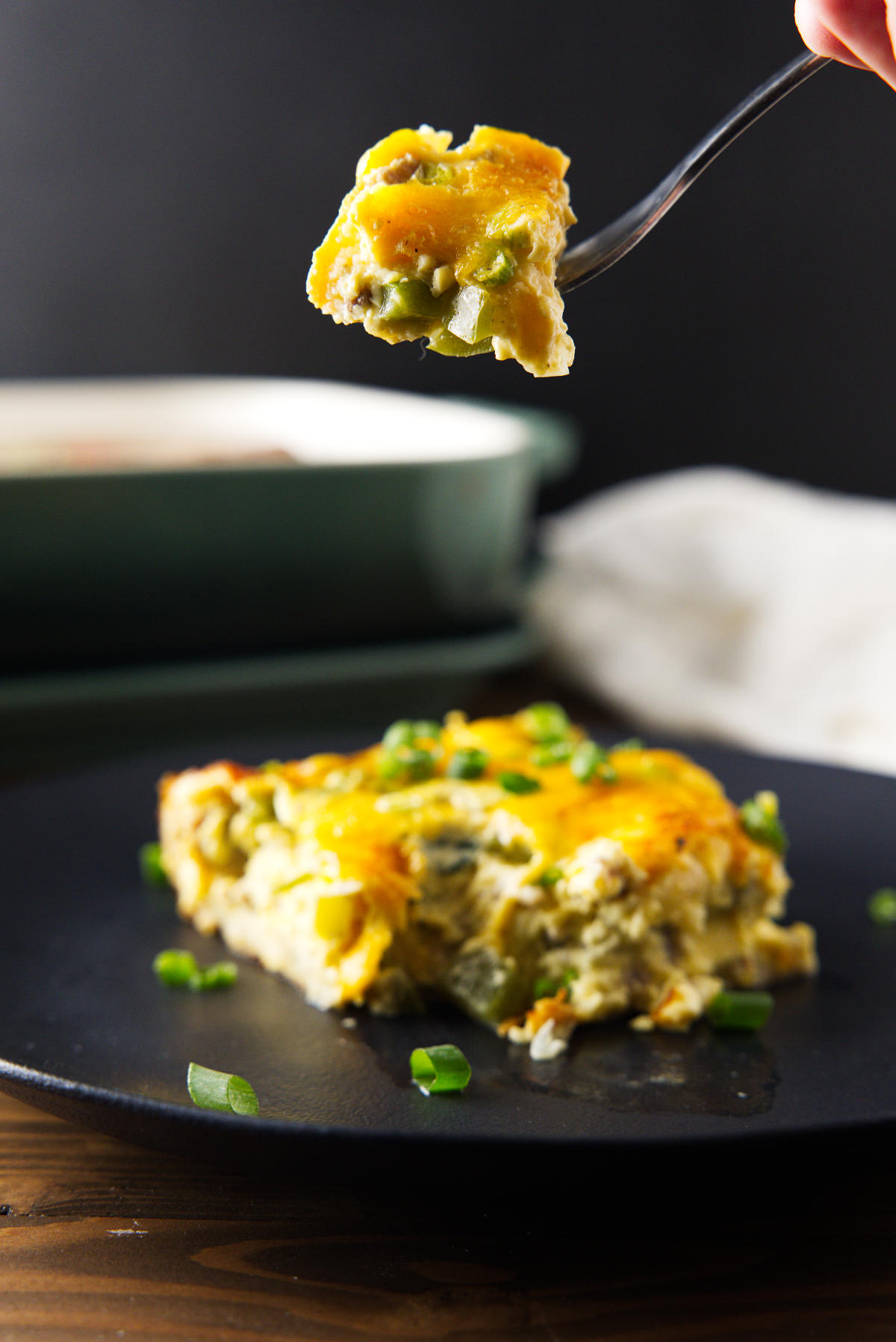 Fork lifting a bite of sausage, egg, and cheese breakfast casserole above a plated slice.