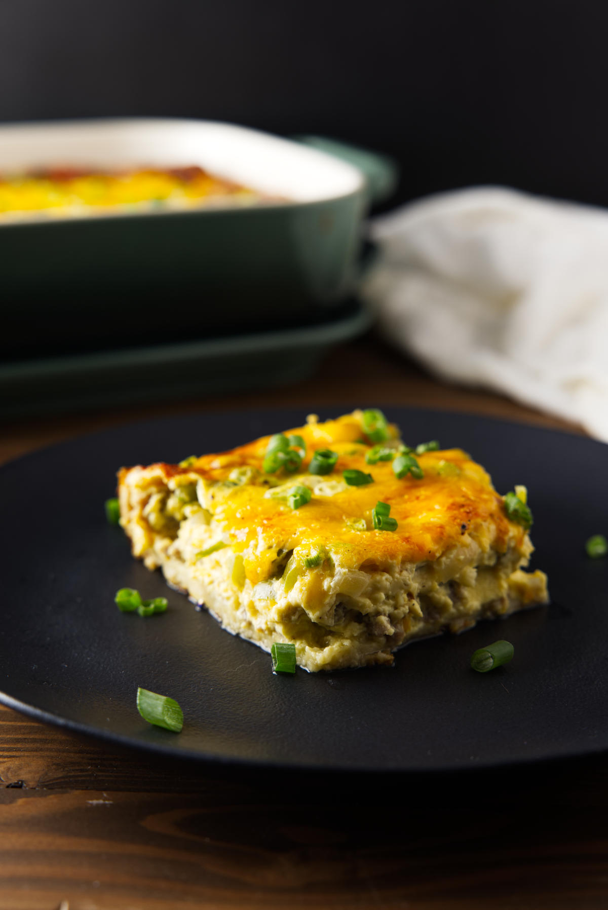 Cheesy breakfast casserole square served on a black plate with the baking dish behind it.
