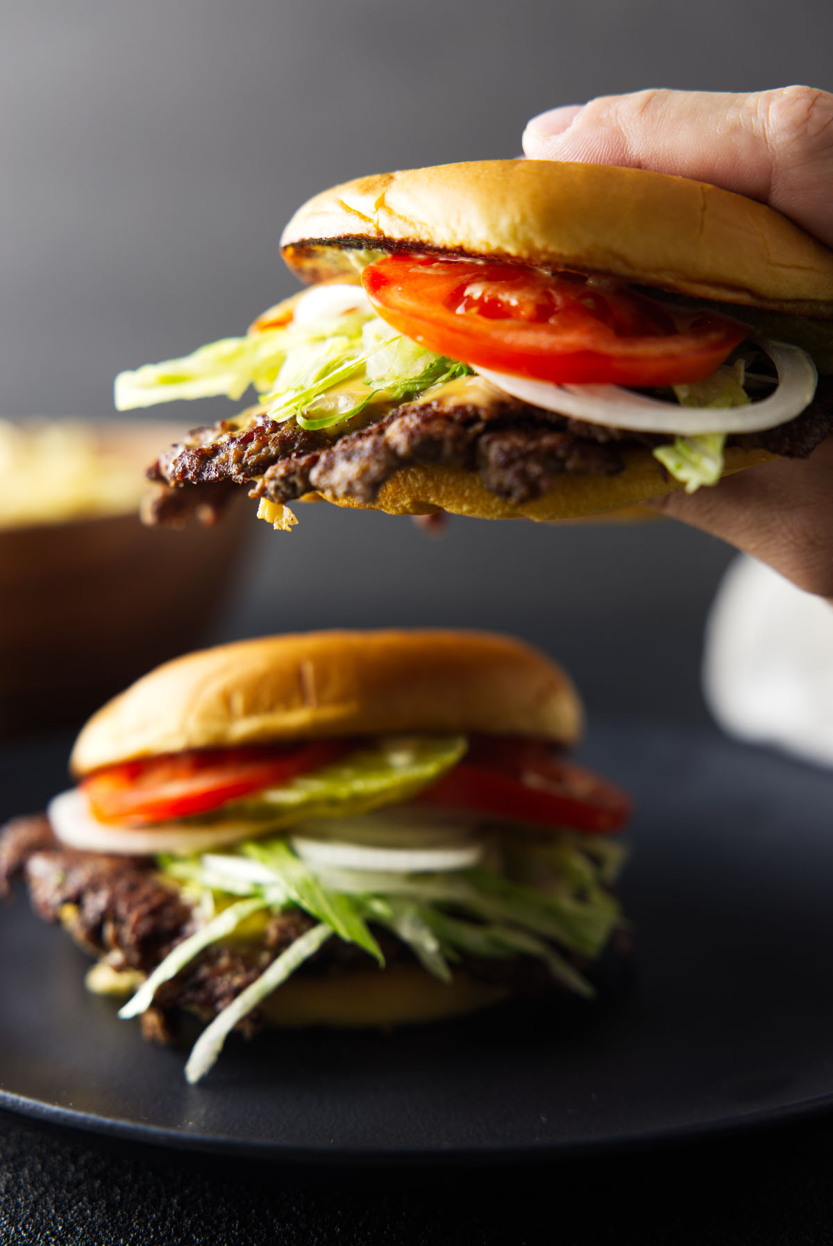 Hand holding a smash burger with tomatoes, pickles, lettuce, and onions loaded on and melted cheese on the burger patty.