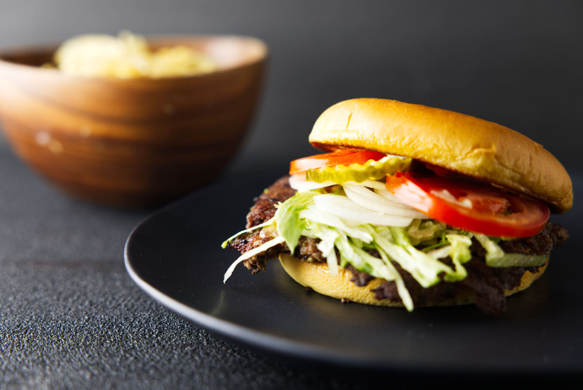 Smash burger loaded with toppings and resting on a black plate with a bowl of potato chips in the background.